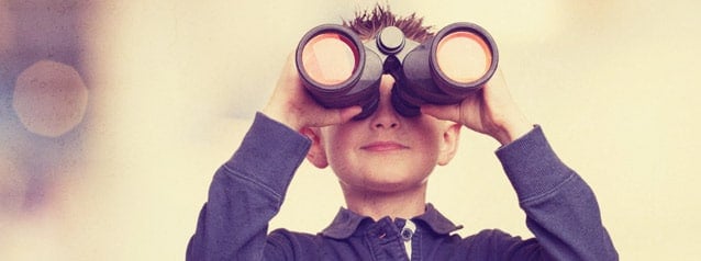 A young boy looking through binoculars at a help desk, focusing intently with a slightly blurred, light background.