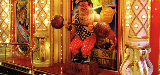 A colorful circus-themed display featuring a caricature statue of a strongman holding barbells, surrounded by decorative lights and vintage posters.