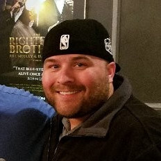 Man in a black cap smiling, standing in front of a movie poster.