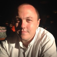 A man with a shaved head, wearing a white shirt, smiling subtly in a dimly lit room.