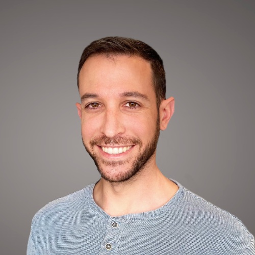 Portrait of a smiling man with short dark hair, wearing a blue shirt, against a gray background.