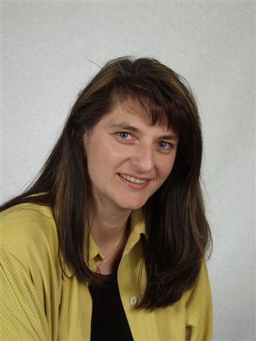 Portrait of a smiling woman with dark hair wearing a yellow cardigan and a black tie, against a plain light background.