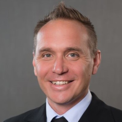 Professional headshot of a smiling man with short, light hair, wearing a dark suit and tie against a gray background.