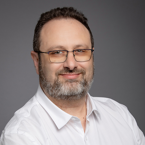 Portrait of a smiling middle-aged man with glasses, wearing a white shirt against a gray background.