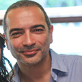 A man with a slight smile looking directly at the camera, featuring a closely shaved head and light stubble. he wears a light blue shirt and is indoors.