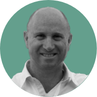 Black and white photo of a smiling bald man in a white shirt, set against a plain, teal background.