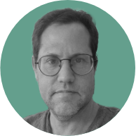 A middle-aged man with glasses and stubble, wearing a gray t-shirt, set against a teal background.