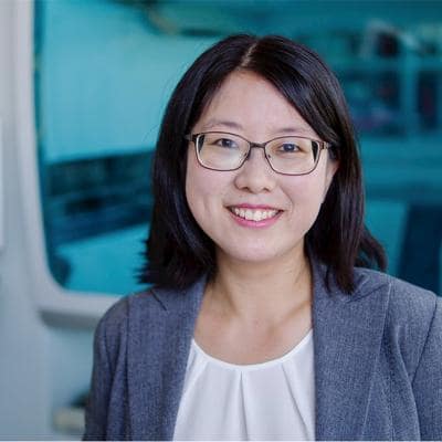 Portrait of a smiling asian woman wearing glasses and a grey blazer over a white blouse, with a blurred blue background.